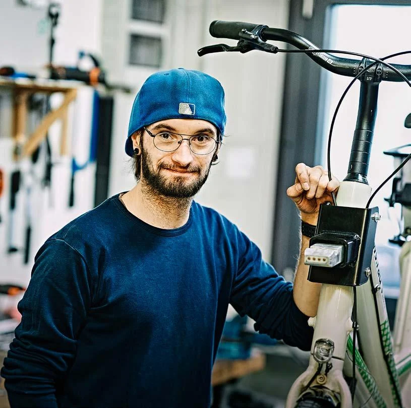 Ein Mann mit Bart, Brille und einer rückwärts getragenen blauen Baseballkappe steht lächelnd neben einem Fahrrad und blickt in die Kamera. Er legt eine Hand locker an die Unterseite des Oberrohrs. Die Person befindet sich links im Bild, während der Fahrradlenker über ihren Kopf hinausragt. Das Fahrrad ist angeschnitten dargestellt. Der Hintergrund ist durch die geringe Tiefenschärfe weich unscharf, sodass der Fokus auf der Person und dem Fahrrad liegt.