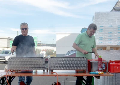 Zwei Männer bereiten an einem Gasgrill auf einem Tisch gemeinsam das Grillgut zu.
