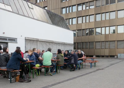 Mehrere Personen sitzen auf Bierbänken vor einer Fabrikhalle und unterhalten sich bei einer Feier.