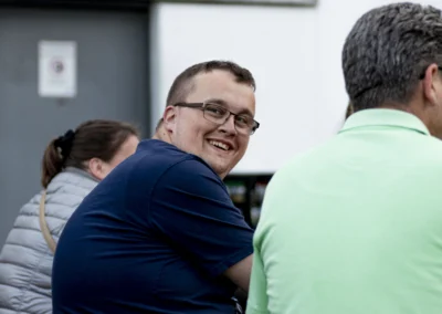 Zwei Männer sitzen nebeneinander; einer lächelt über die Schulter in die Kamera.