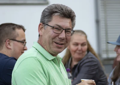 Sitzender Mann mit grauen Haaren und Brille im grünen Poloshirt dreht sich lächelnd zur Kamera um.